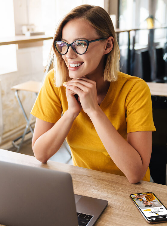 Woman in yellow shirt smiles while at her computer and her phone with the Cornhusker Bank website open