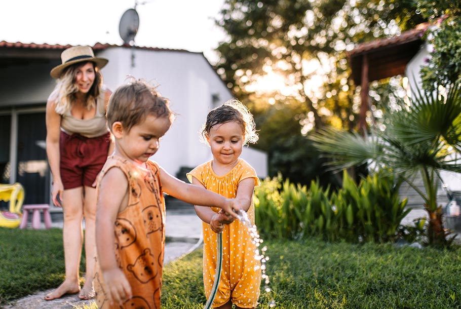 Mother watches her two little ones playing with a hose spraying water